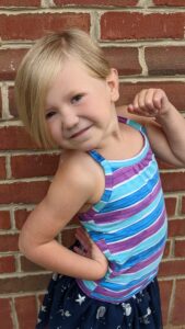 A small white girl shows off her new blond bob by posing to the side, with one hand on her hip and the other pointing at her face. She wears a blie and purple striped tank top.