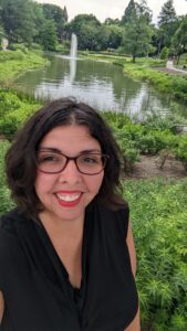 A 30something year old Latina woman with a dark wavy bob, red glasses, red lipstick, and a black blouse smiles at the camera. Greenery and a small pond are behind her.