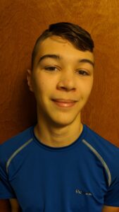 A young, white teenage boy smirks at the camera. He's standing in front of a door, wearing a dark blue athletic shirt and has close cropped brown hair that's shaved on the sides.